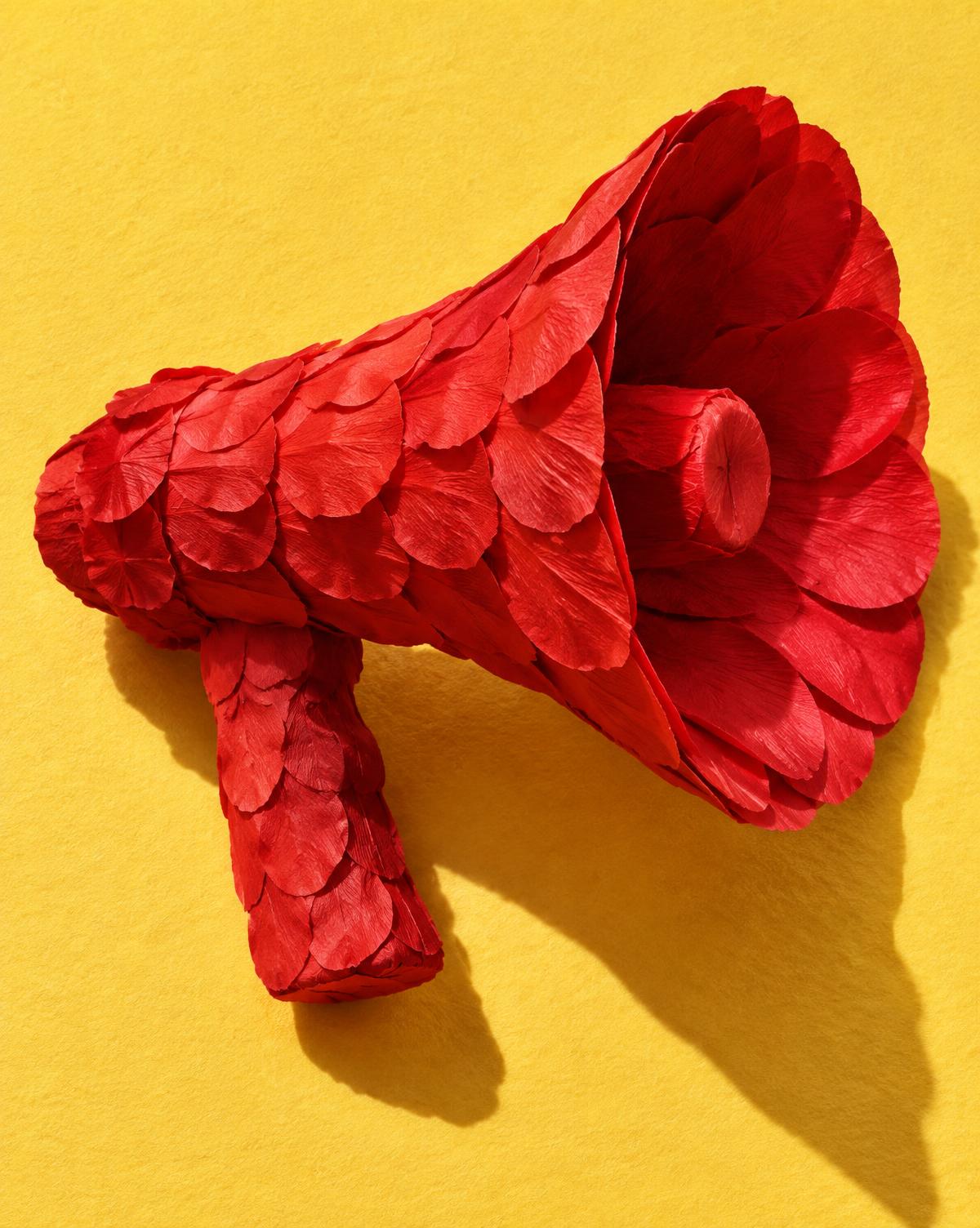A megaphone built from folded red paper petals on a butter yellow backdrop